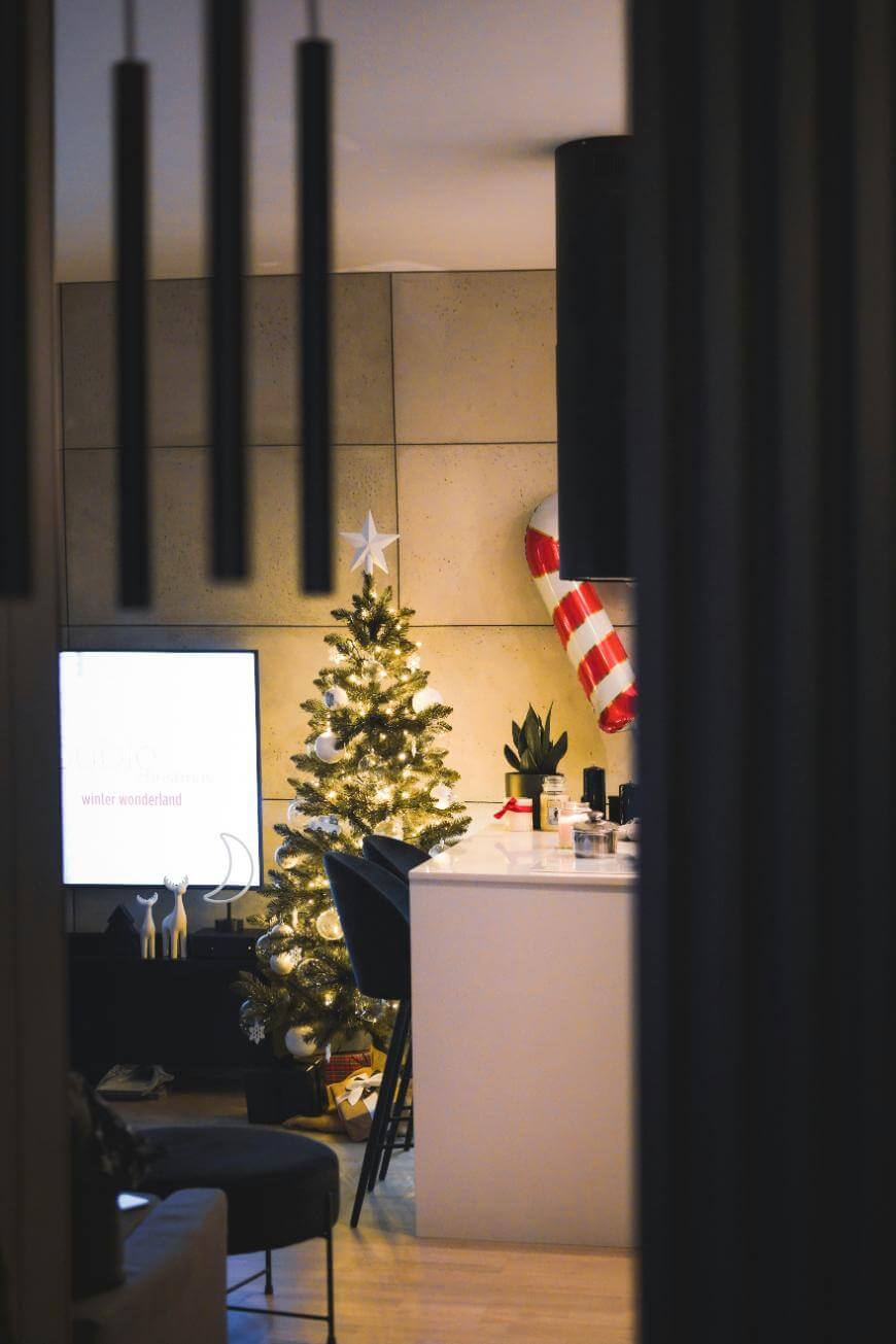 A cozy, modern living room decorated for Christmas with a lit tree beside a TV, a large candy cane decoration, and holiday ornaments on a white kitchen island. Warm lighting creates a festive atmosphere.