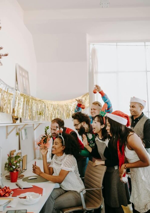 A group of people in festive attire and Santa hats happily gather around a woman at a desk in a decorated office, celebrating and looking at a computer screen together.