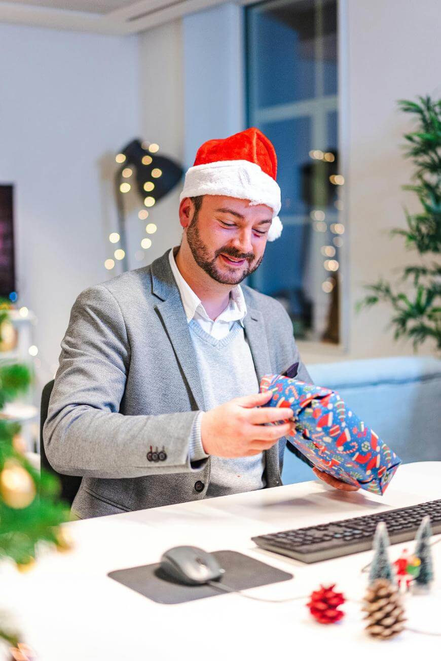 A man in a gray blazer and Santa hat sits at a desk, smiling as he opens a wrapped gift. The office is decorated with Christmas lights and a tree, creating a festive atmosphere.