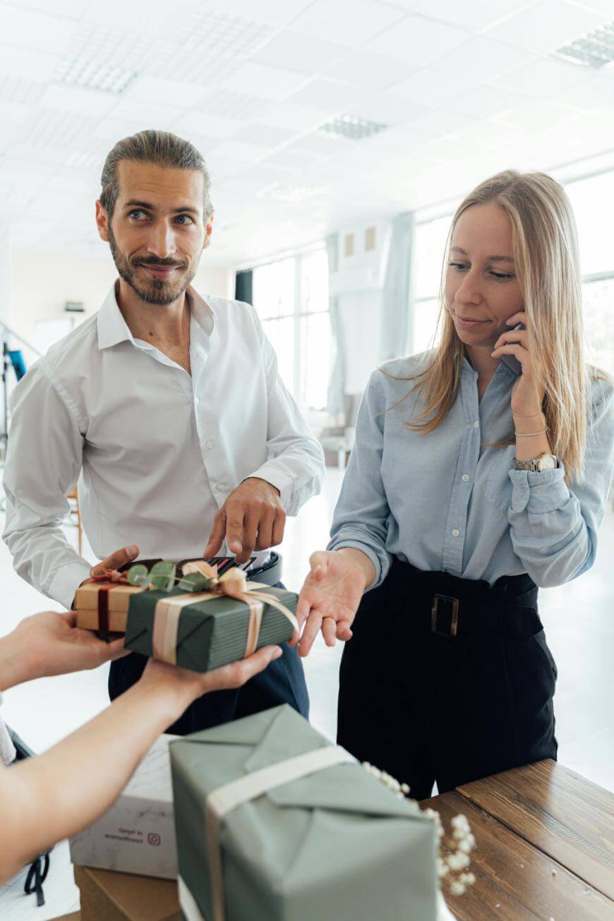 A man and woman in business attire stand near a table, smiling as a person hands them a wrapped gift. The room is bright with large windows and a modern, professional atmosphere.