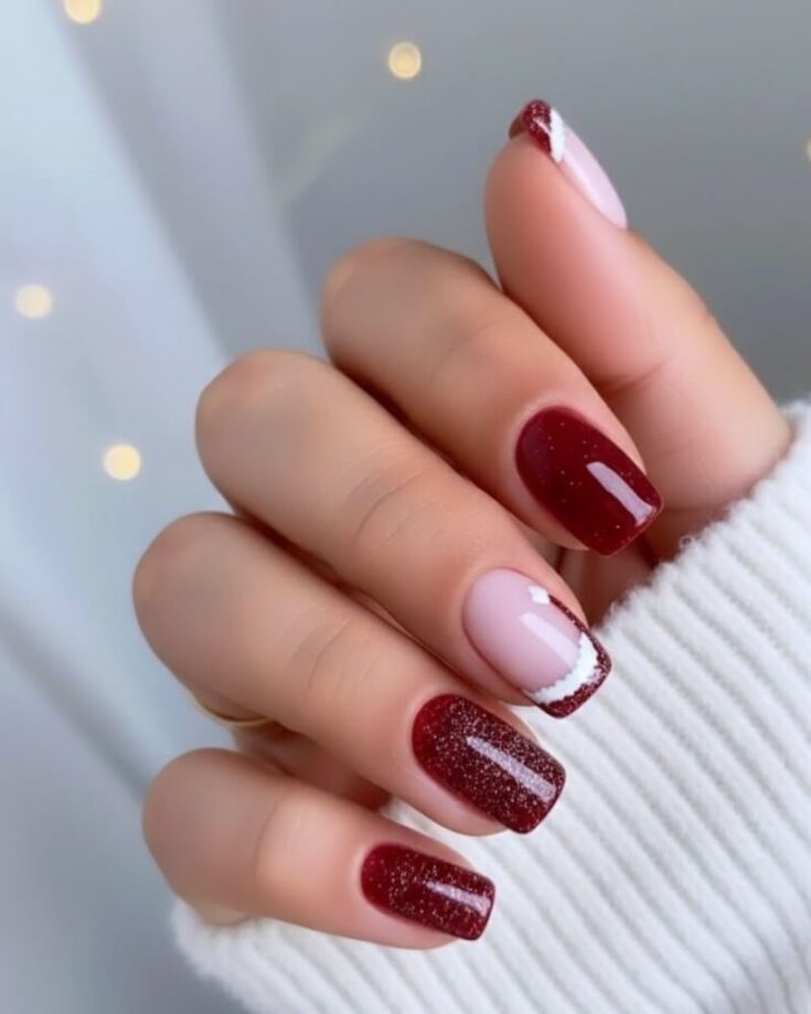 A hand with manicured nails featuring a mix of deep red glitter polish and light pink polish with red and white diagonal accents. The person is wearing a white ribbed sweater. Soft lights blur in the background.
