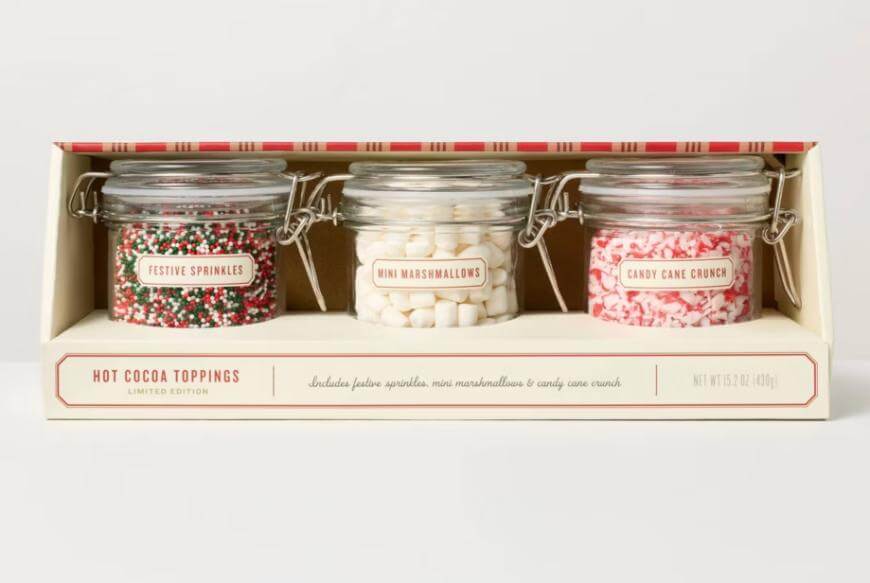 A boxed set of three glass jars labeled “Festive Sprinkles,” “Mini Marshmallows,” and “Candy Cane Crunch.” The jars contain holiday-themed hot cocoa toppings and are arranged side by side in festive packaging.