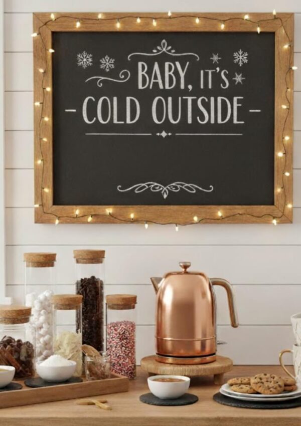 A cozy kitchen setup with jars of toppings, a copper kettle, mugs, and a chalkboard sign framed in wood and string lights reading Baby, its cold outside.