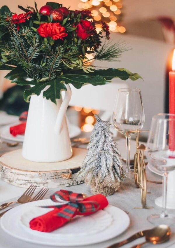 A festive table setting with a white pitcher filled with red flowers and greenery, a small frosted decorative tree, red napkins tied with plaid ribbon, wine glasses, and a lit red candle. Warm lights glow in the background.