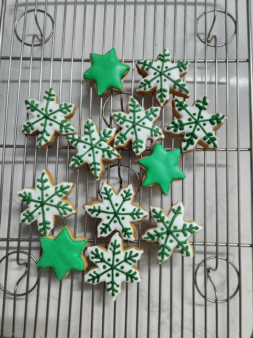Decorated cookies on a cooling rack, shaped like stars and snowflakes. The snowflake cookies are white with green icing details, while the star cookies are frosted solid green. The rack sits on a marble surface.