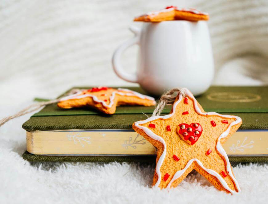 Star-shaped cookies decorated with white and red icing rest on a closed green book, with one cookie hanging by a string. A white mug and another cookie are in the blurry background on a soft, white surface.