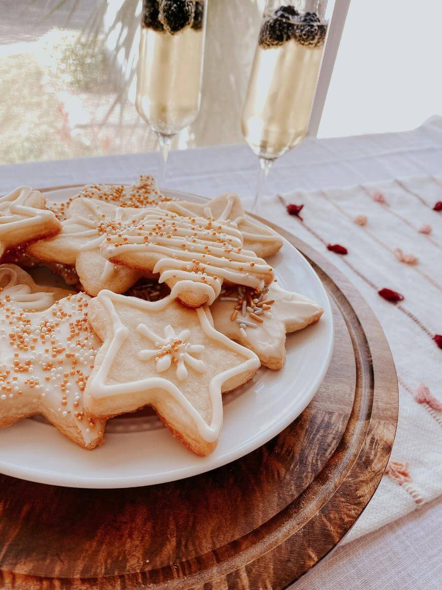 A plate of iced star-shaped sugar cookies with sprinkles sits on a wooden tray next to two glasses of sparkling drink garnished with blackberries, on a light-patterned tablecloth by a sunny window.