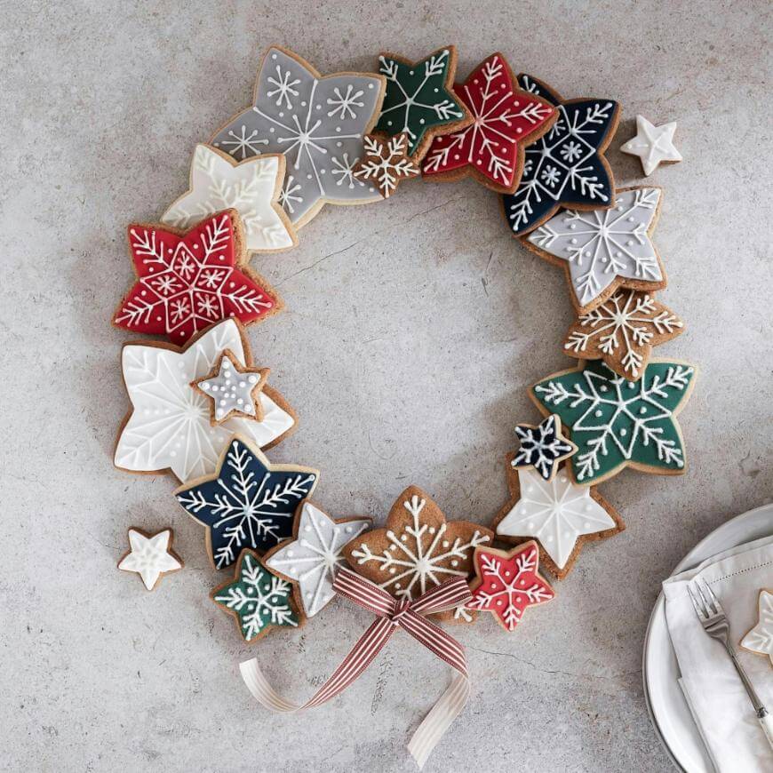 A festive wreath made of star and snowflake-shaped cookies in red, green, white, and gray icing, arranged in a circle on a light surface with a striped ribbon bow and a plate with cutlery nearby.