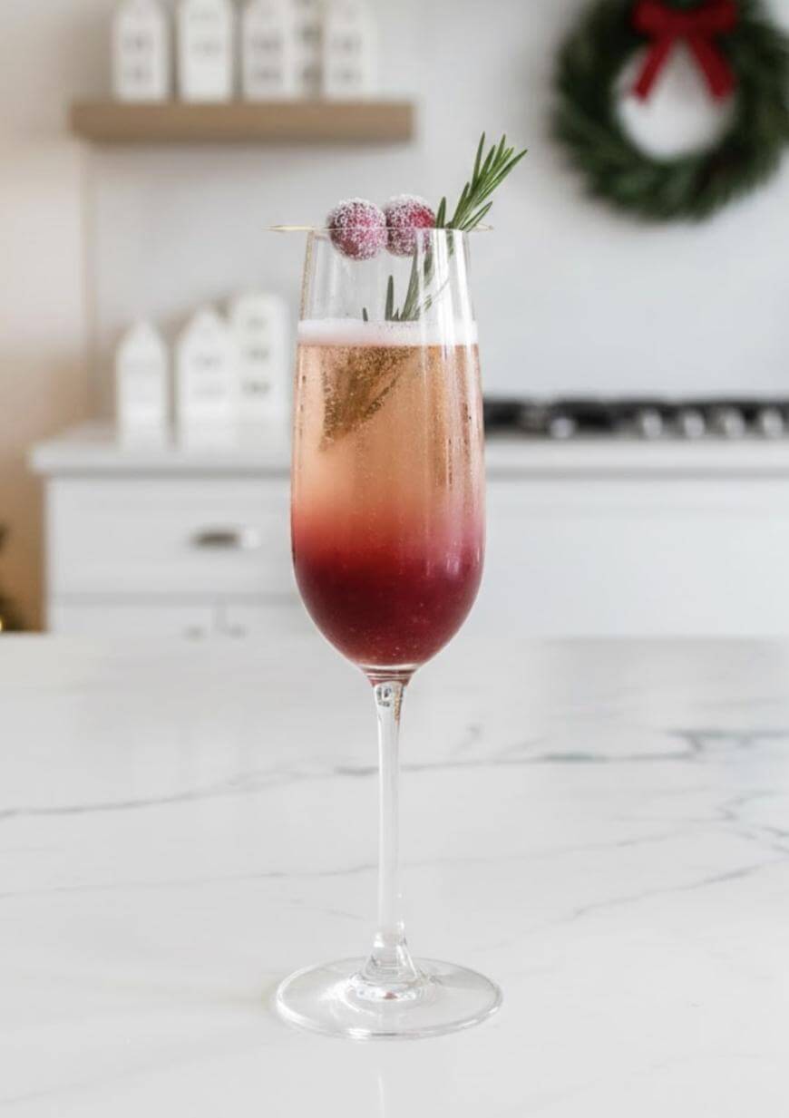 A tall champagne flute filled with a layered red and pink drink, garnished with sugared cranberries and a sprig of rosemary, sits on a marble countertop in a bright kitchen decorated for the holidays.