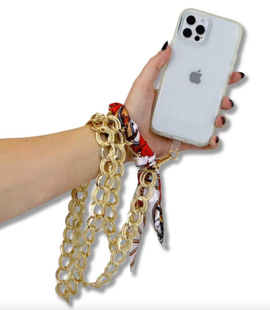 A hand holds an iPhone in a clear case with a gold chain strap attached. The chain is wrapped around the wrist and decorated with a red, white, and brown patterned scarf.