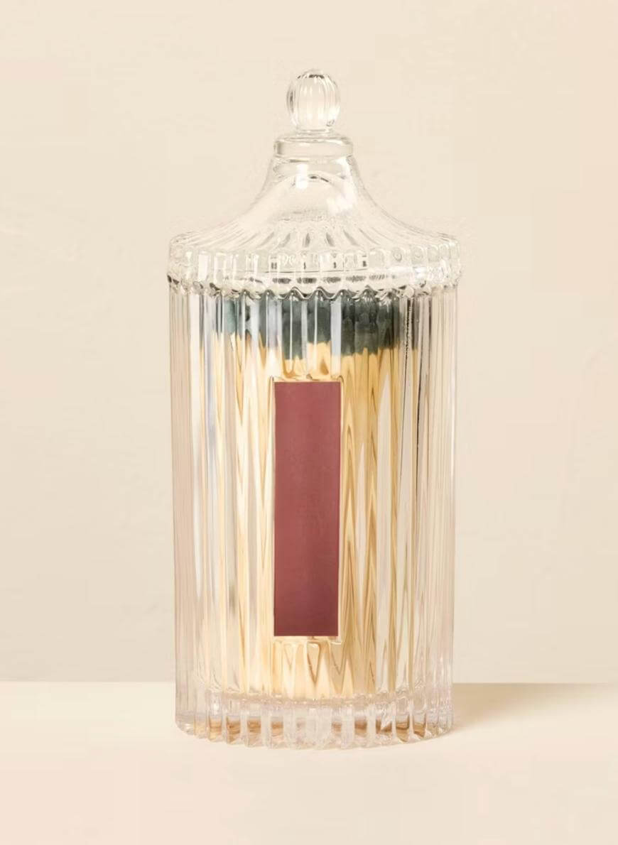 A tall, clear, ribbed glass jar with a lid, containing a candle. The jar has a rectangular maroon label on the front and is set against a plain, light background.