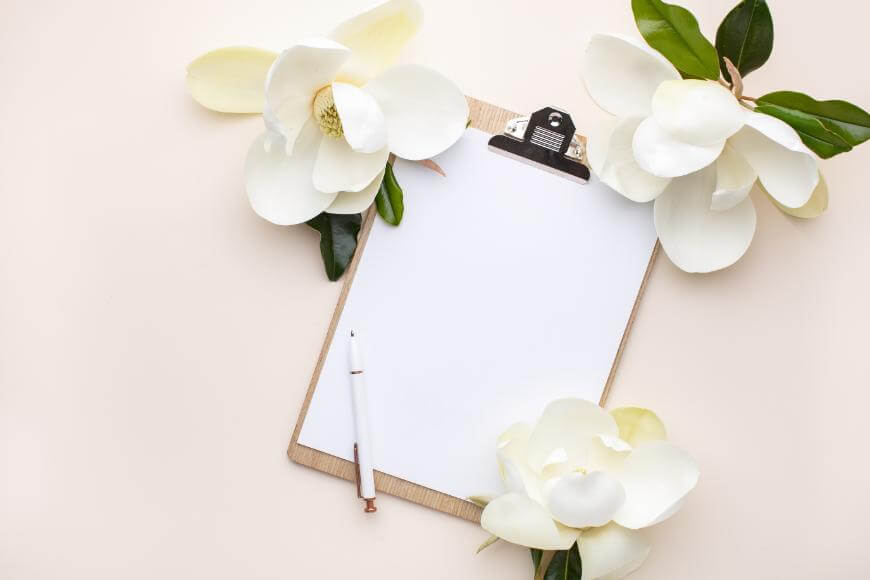 A blank clipboard with a white sheet of paper and pen is surrounded by large white magnolia flowers and green leaves on a light beige background.