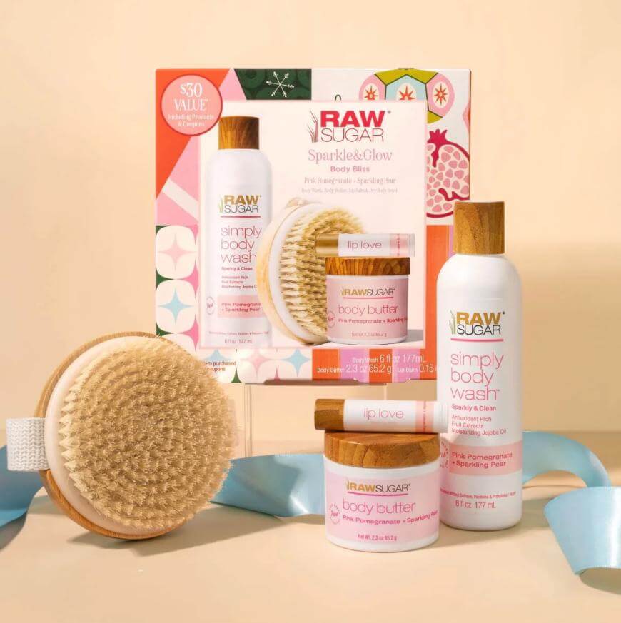 A Raw Sugar Sparkle & Glow Body Bliss gift set with body wash, body butter, a wooden dry brush, and their packaging. All products feature light pink and wood accents and are displayed against a neutral background.