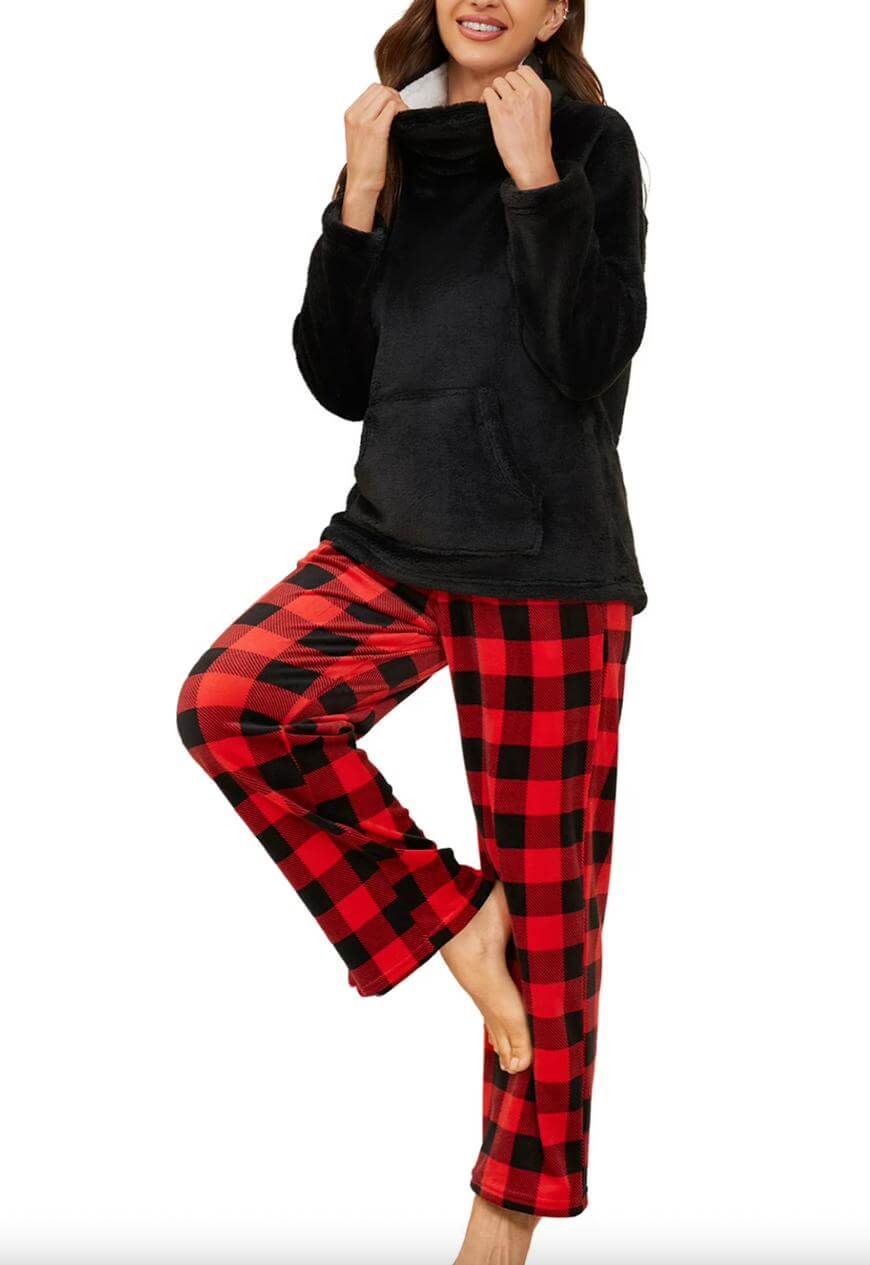 A woman stands barefoot, wearing a black fleece pullover and red-and-black buffalo plaid pajama pants. She has one knee raised and holds the collar of her top, smiling. The background is white.
