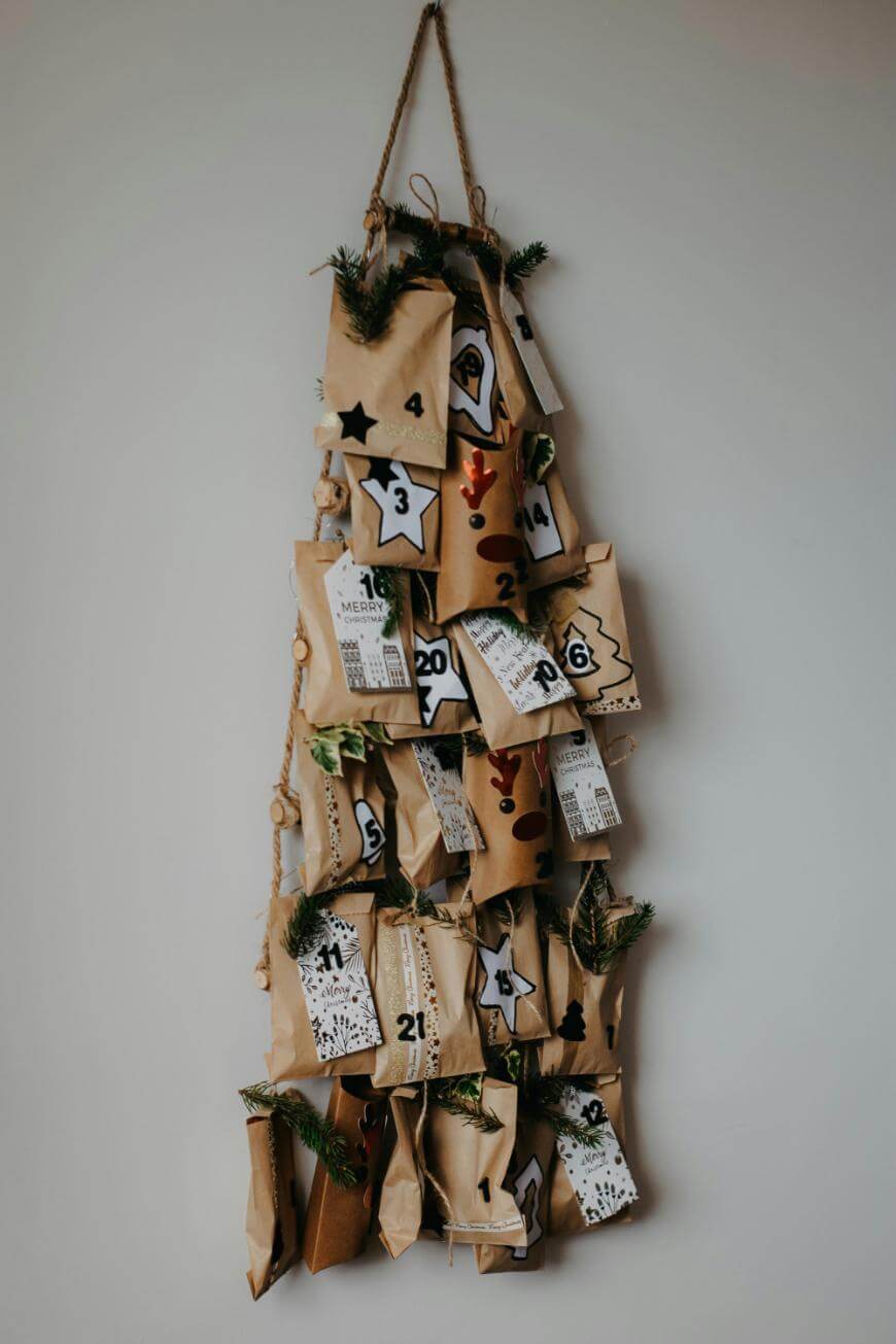 A homemade advent calendar shaped like a Christmas tree, made of brown paper bags, each labeled with numbers and decorated with greenery, hanging on a wall with twine.