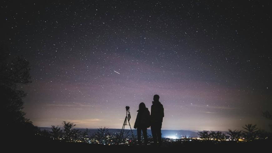 Two people stand beside a tripod, looking up at a star-filled night sky with a faint shooting star visible above a distant city’s lights on the horizon. The scene suggests stargazing or astronomy.