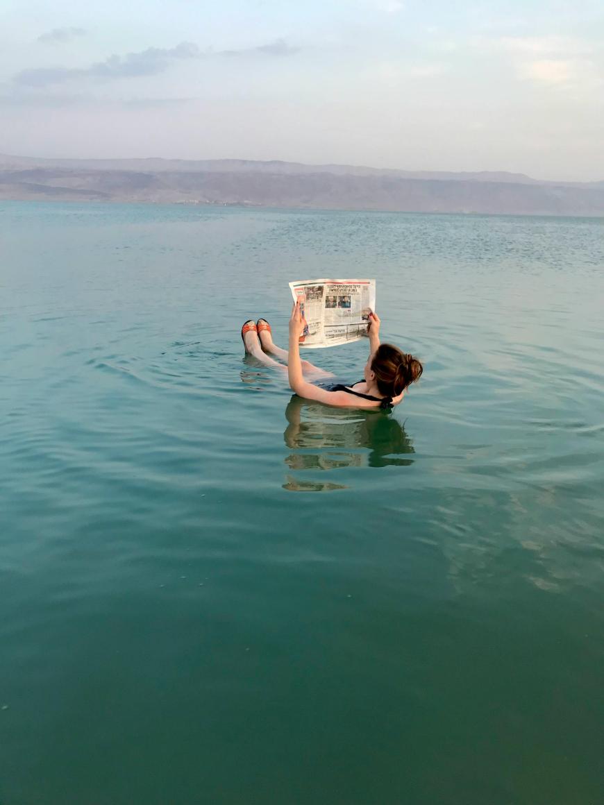 A person in a swimsuit floats on their back in calm blue water, reading a newspaper. The setting appears tranquil with distant mountains and a cloudy sky in the background.