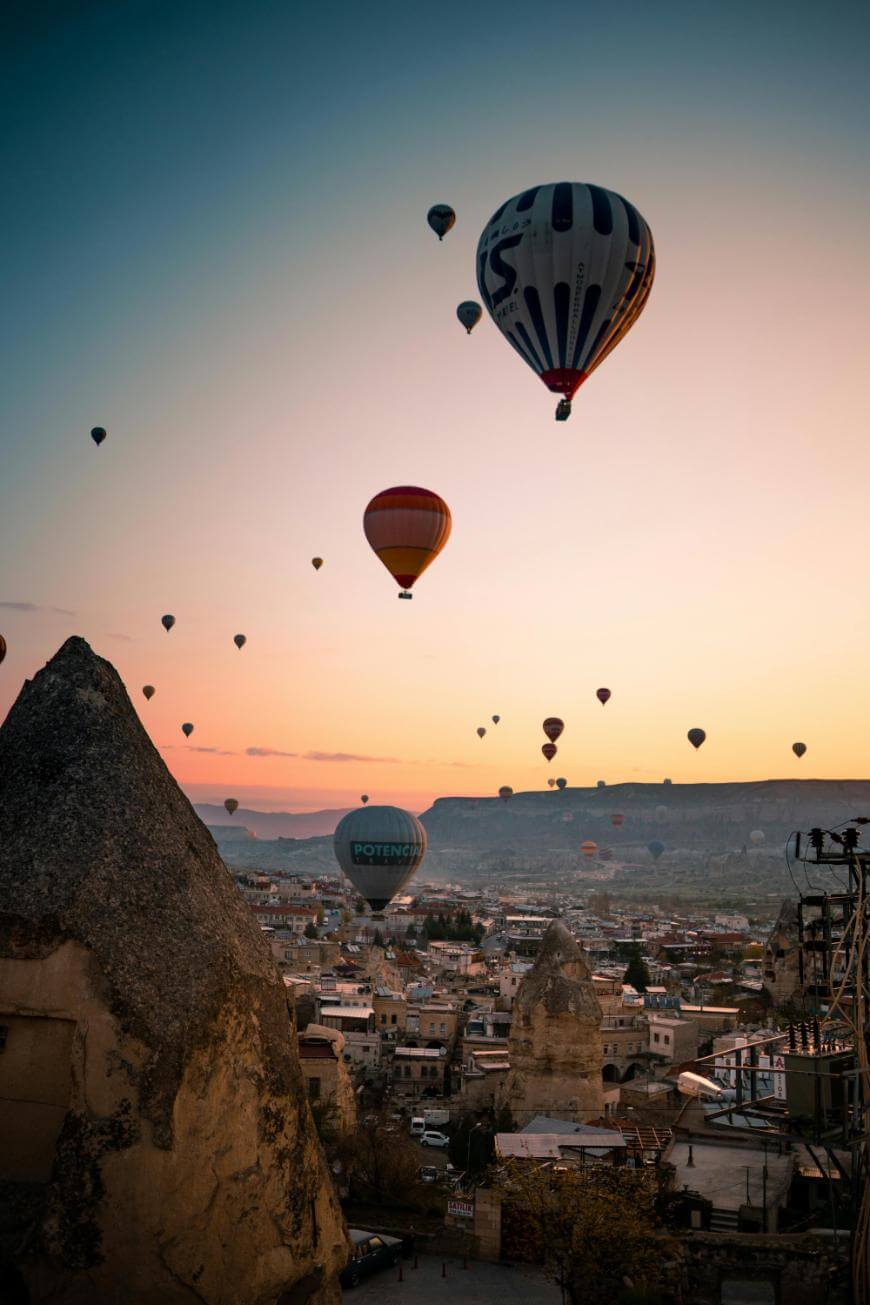 Hot air balloons float above a town with rocky formations at sunrise or sunset, casting a warm light over the landscape, likely in Cappadocia, Turkey.