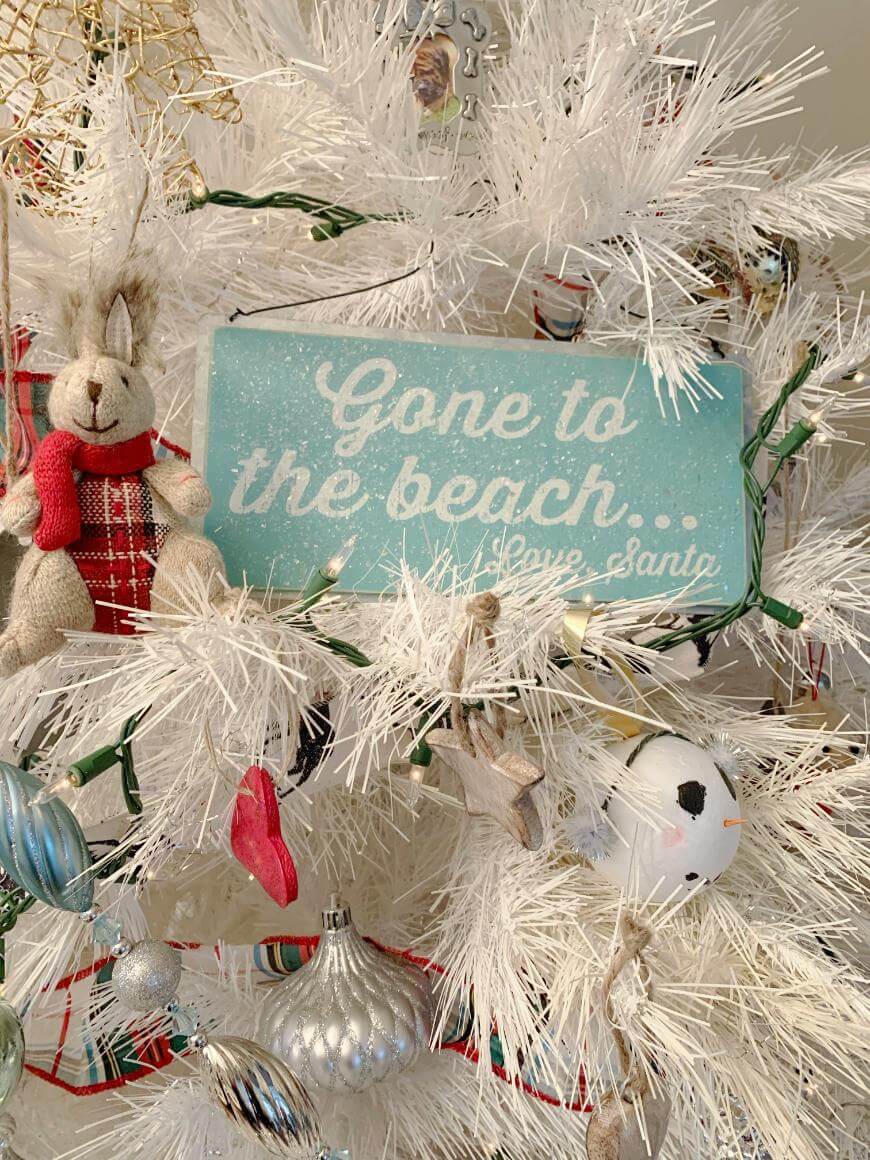 A white Christmas tree decorated with ornaments, including a bunny, a soccer ball, and a blue sign that reads “Gone to the beach… Love, Santa,” surrounded by white branches and holiday decorations.
