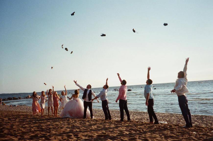 A group of people dressed in formal attire stand on a sandy beach, joyfully throwing their hats into the air with the ocean and clear sky in the background.