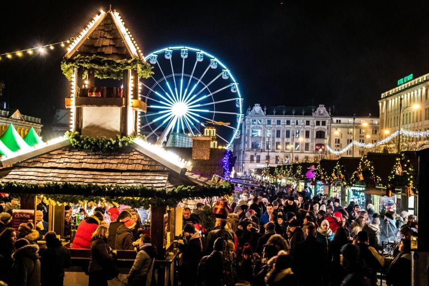 A festive Christmas market at night, crowded with people. Bright lights decorate stalls and a large Ferris wheel glows in the background, with historic buildings also illuminated.