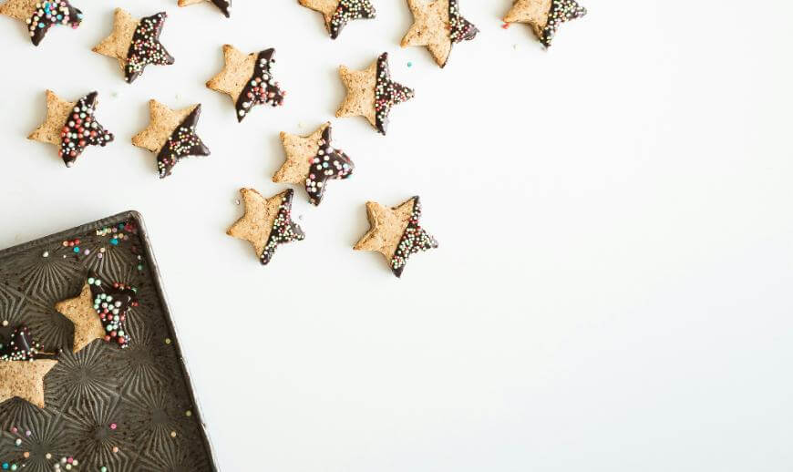 Star-shaped cookies dipped in chocolate and topped with colorful sprinkles are scattered on a white surface, with some cookies on a textured baking tray in the bottom left corner.