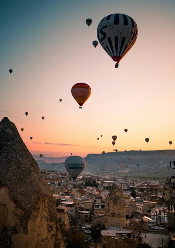 Hot air balloons float above a rocky townscape at sunrise or sunset, with warm light illuminating the sky and unique rock formations in the foreground, likely in Cappadocia, Turkey.