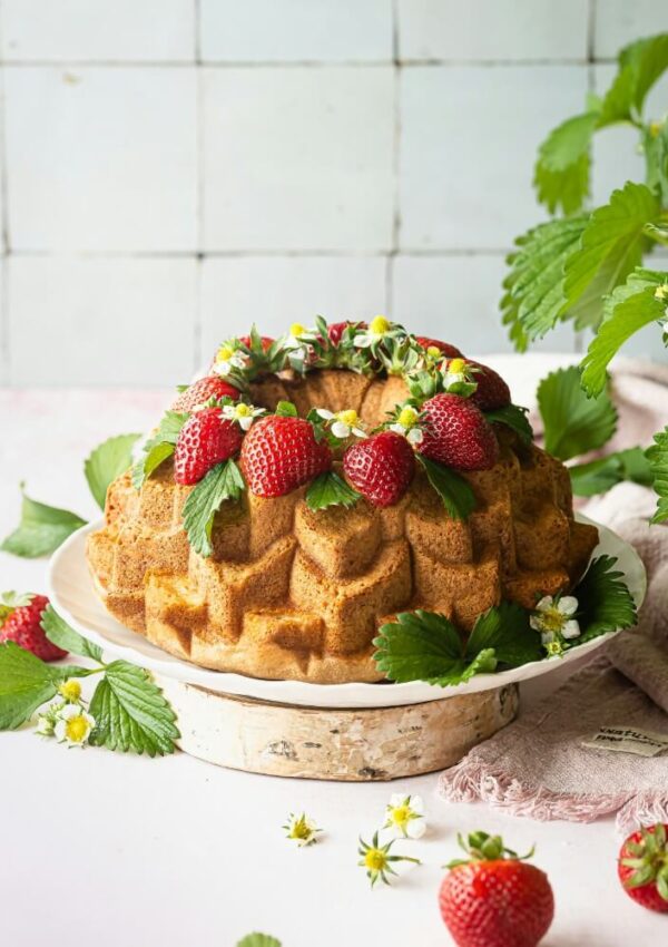 A bundt cake on a white plate is decorated with fresh strawberries, green leaves, and small white flowers. More strawberries and leaves are scattered around, with a plant visible on the right.