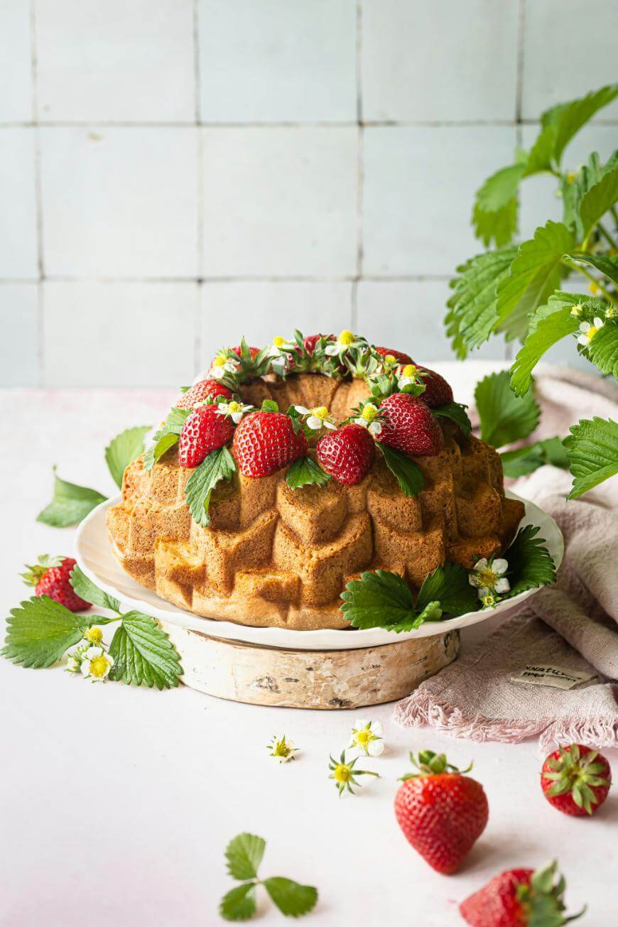 A decorative bundt cake topped with fresh strawberries, green leaves, and small white flowers sits on a plate, with more strawberries, leaves, and flowers scattered around on a light surface.