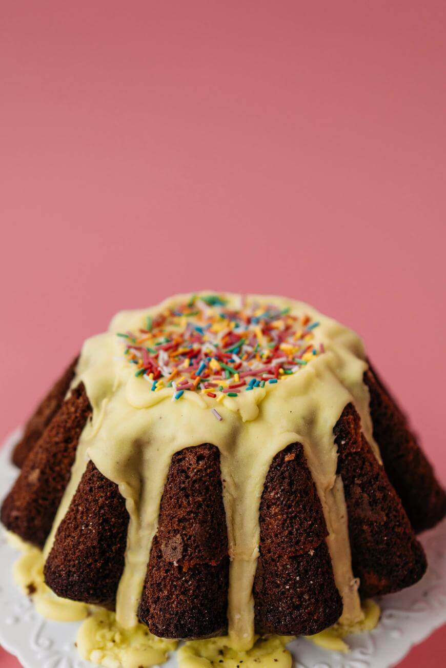 A chocolate bundt cake with yellow icing dripping down the sides and colorful sprinkles on top, displayed on a white cake stand against a pink background.