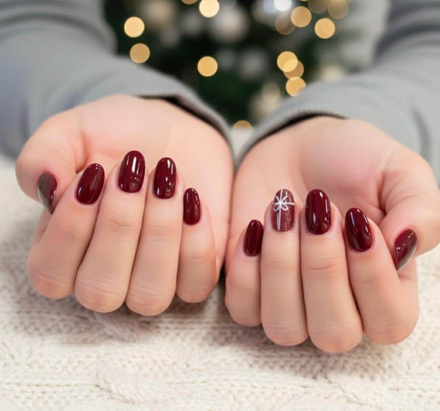 Close-up of hands with dark red manicured nails, one nail featuring a white snowflake design on a shimmering accent nail. Background shows blurred festive lights and a hint of a Christmas tree.