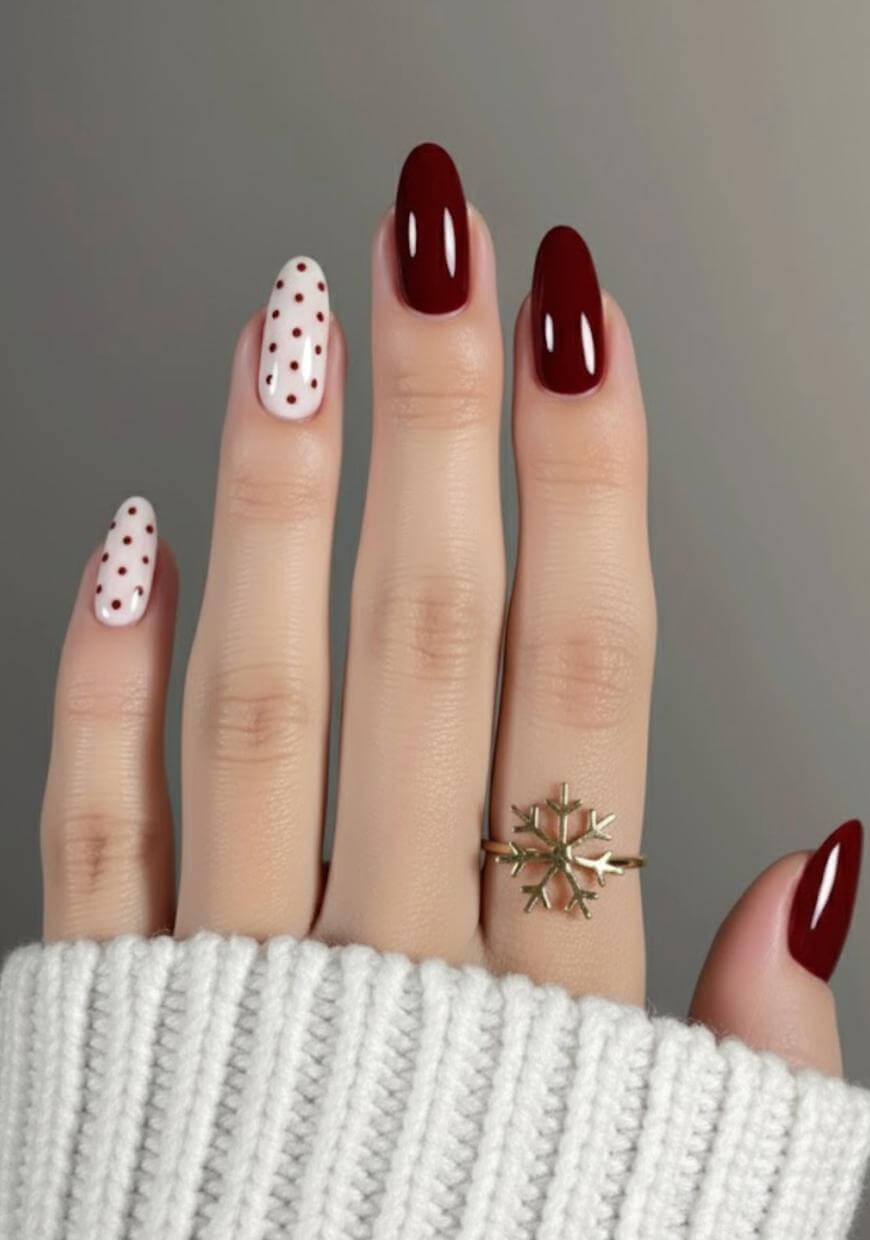 A hand with manicured nails; three nails are painted dark red, while the index and ring nails are white with small red polka dots. The person wears a gold ring and a white knitted sleeve covers the wrist.