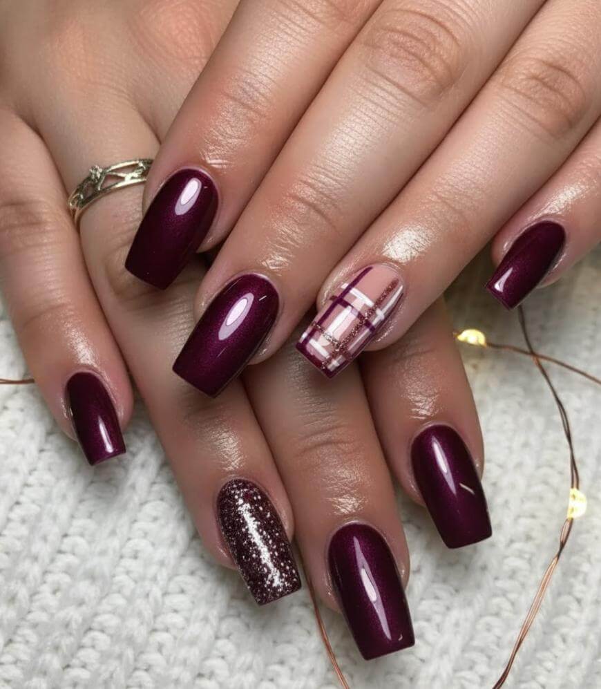 Hands with square-shaped nails painted in shiny burgundy polish; one accent nail features a plaid design in burgundy and gold on a nude base, and another has silver glitter. Hands rest on a white knitted surface.