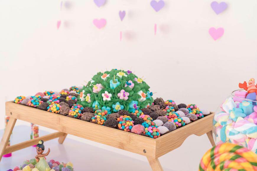 A colorful candy display on a wooden tray features clusters of various sweets, including multicolored round candies, chocolate pieces, and flower-shaped candies, with heart decorations hanging in the background.