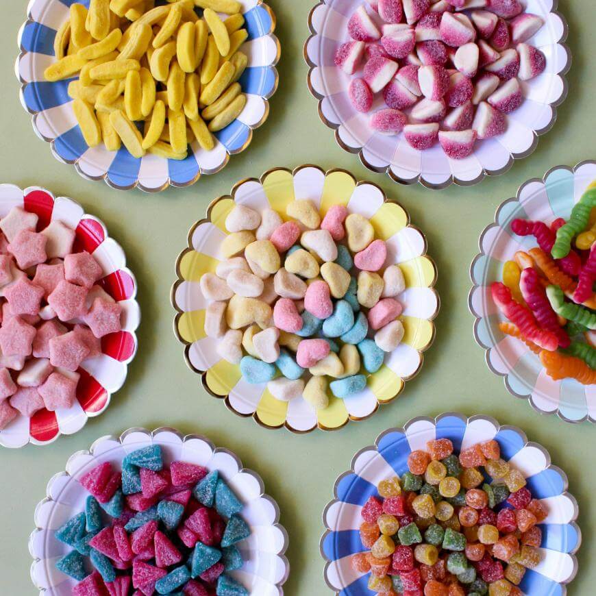 Six colorful plates arranged in a circle, each filled with different types of assorted gummy candies in various shapes and colors, on a light green surface.