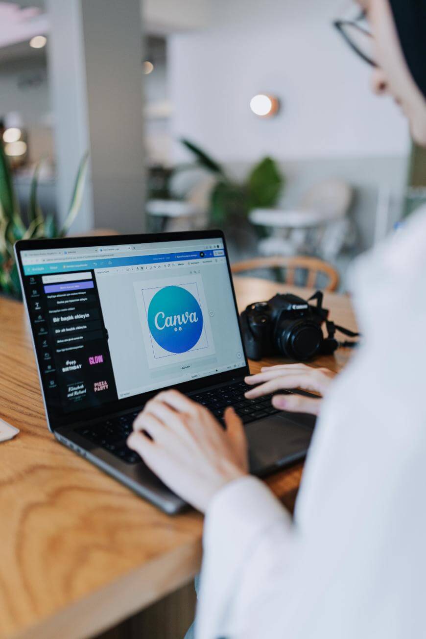 A person works on a laptop at a wooden table, editing a Canva project with a blue and purple circle logo on the screen. A digital camera sits beside the laptop, and the background is a bright café.
