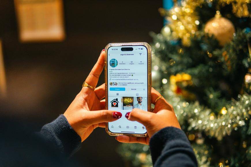 A person holding a smartphone and browsing a social media profile next to a decorated Christmas tree. The persons nails are painted red, and they are wearing rings and a dark sweater.