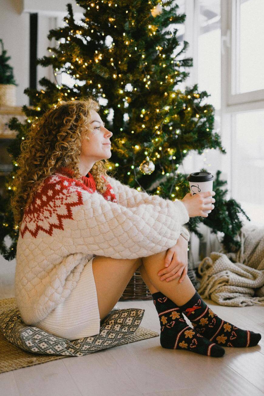 A woman with curly hair wearing a festive sweater and patterned socks sits on a cushion by a decorated Christmas tree, holding a takeaway coffee cup and looking contentedly out a bright window.