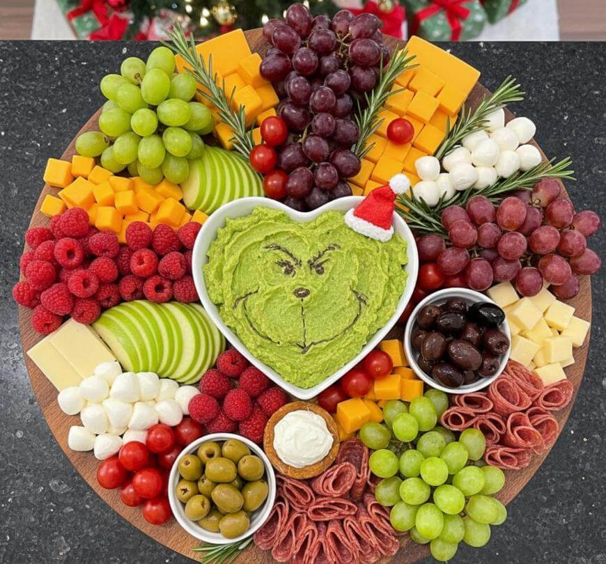 A festive charcuterie board with grapes, cheese cubes, apple slices, tomatoes, olives, raspberries, mozzarella balls, meats, and a bowl of guacamole shaped like the Grinch, wearing a small Santa hat, in the center.
