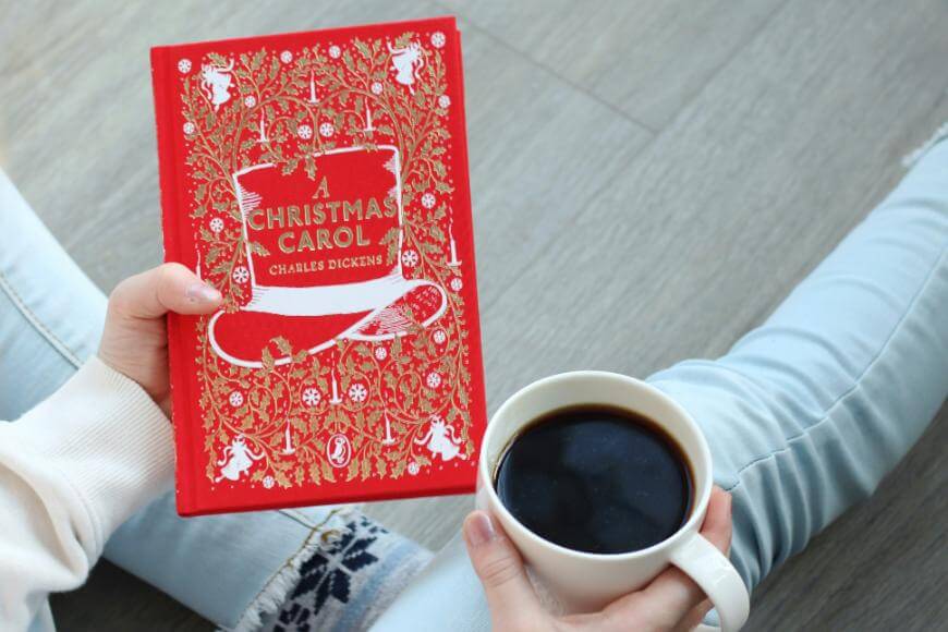 A person sits on the floor holding a red, decorative copy of A Christmas Carol by Charles Dickens in one hand and a white mug filled with black coffee in the other.