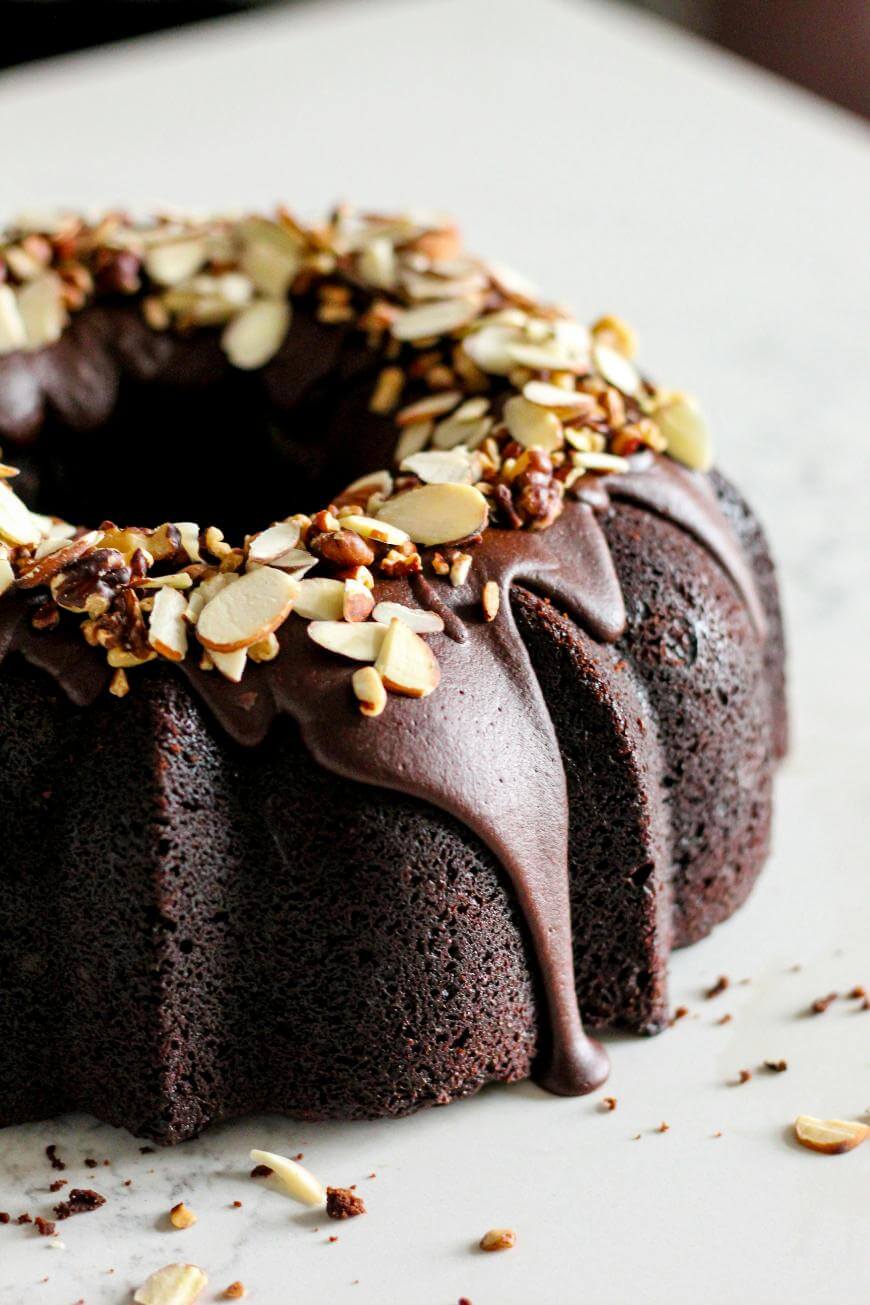 A chocolate bundt cake topped with glossy chocolate glaze, sliced almonds, and chopped nuts, resting on a light-colored surface.