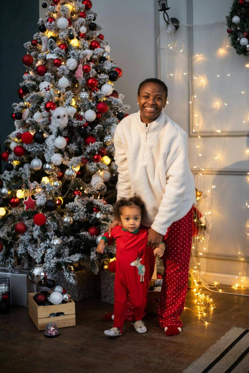 A smiling adult and a toddler in festive pajamas stand by a decorated Christmas tree with red, gold, and white ornaments, surrounded by string lights and presents.