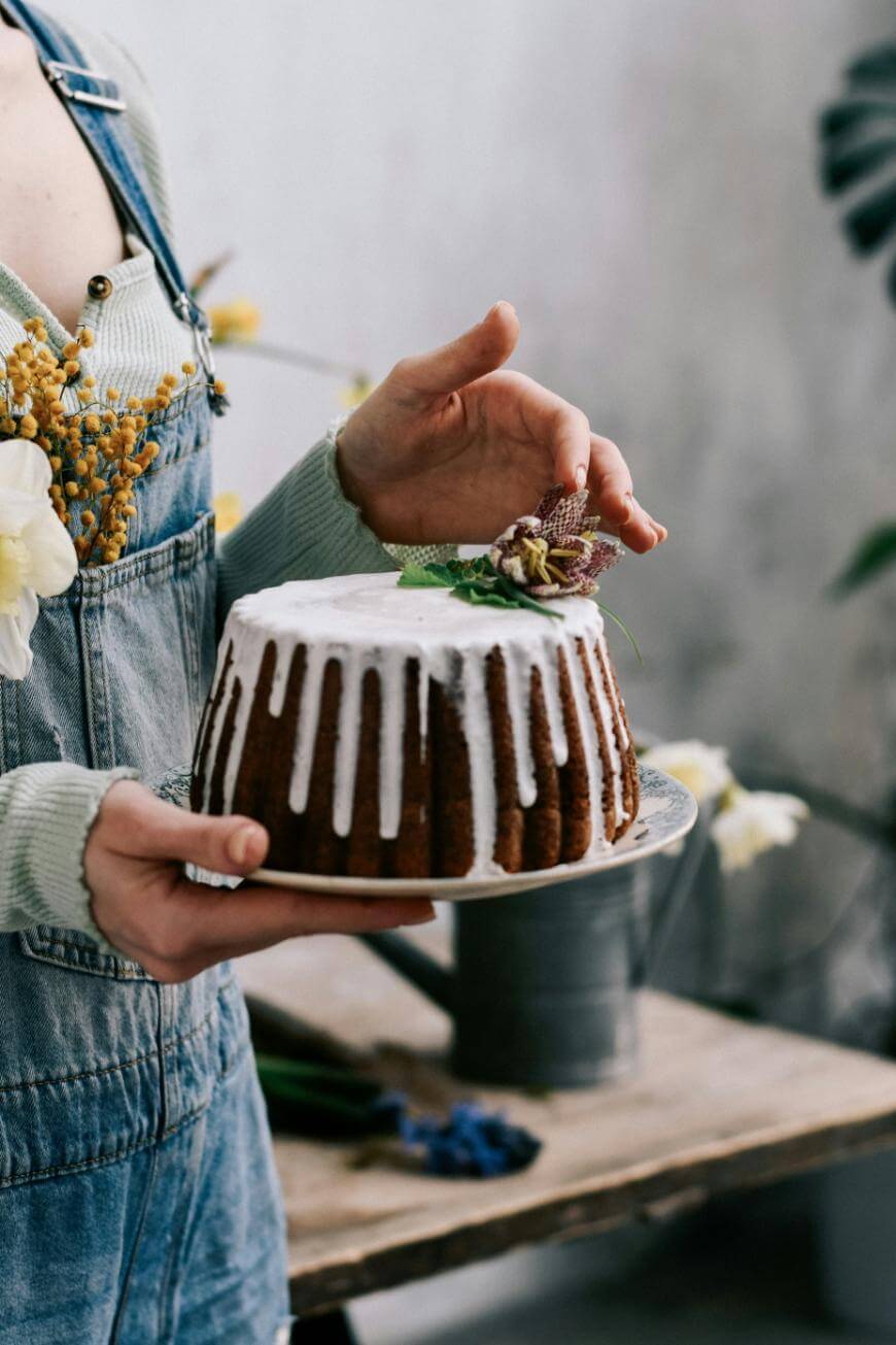 A person in a light sweater and denim overalls holds a round bundt cake with white icing, gently touching a decorative flower and mint leaves on top. Flowers are tucked into their overalls, and plants are in the background.