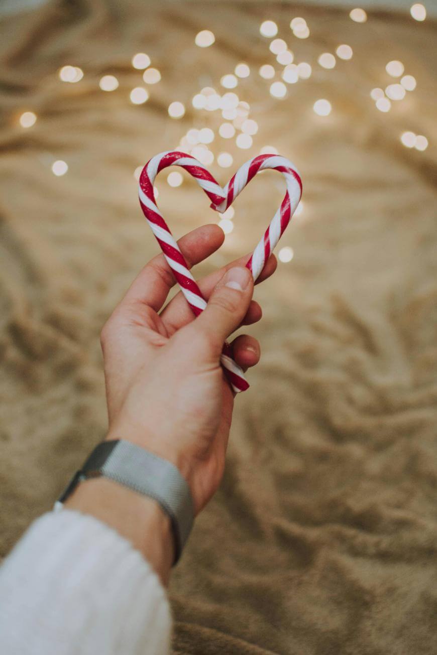 A hand holding two red and white candy canes forming a heart shape, with warm fairy lights softly glowing in the blurred background over a beige, textured surface.