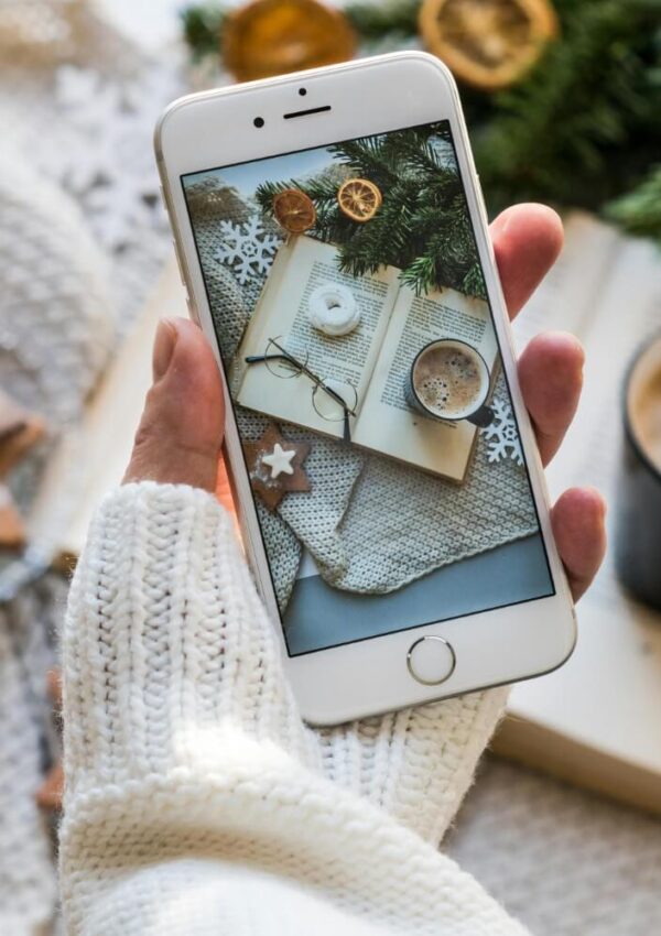 A hand in a white knit sweater holds a smartphone showing a photo of a cozy scene with an open book, a cup of coffee, cookies, and pine branches on a knitted blanket. A coffee cup is blurred in the background.