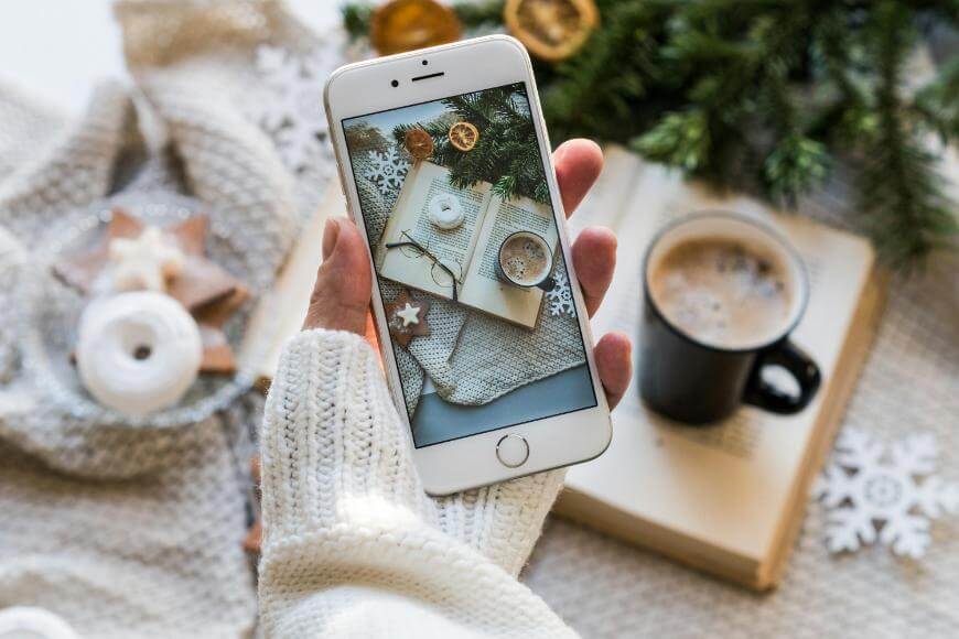 A hand in a white knit sweater holds a smartphone, taking a photo of a cozy scene with a cup of coffee, a book, Christmas cookies, and festive decorations on a knit blanket.