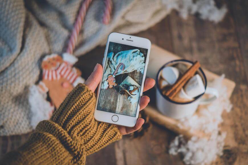 A hand in a mustard knit glove holding a phone, taking a photo of a mug with marshmallows and cinnamon, a gingerbread man, and festive holiday items on a wooden table.
