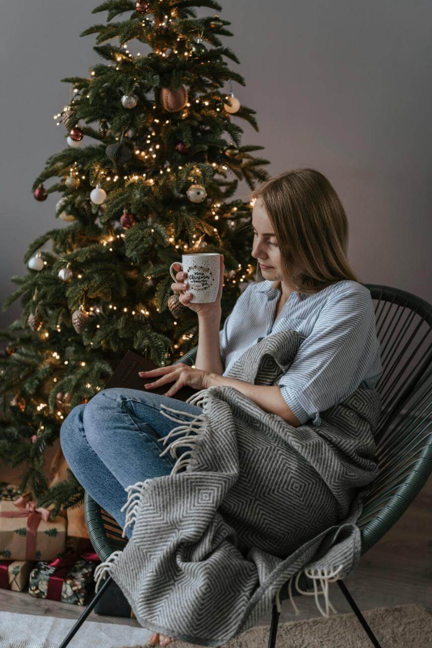A woman sits in a chair by a decorated Christmas tree, wrapped in a gray blanket, holding a mug and reading a book. Wrapped gifts are under the tree and warm lights create a cozy atmosphere.
