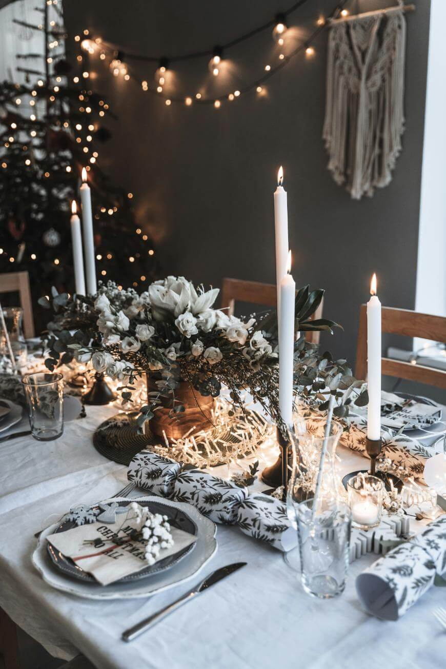 A festive dining table is set with white plates, decorative napkins, white candles, greenery, and string lights. In the background, a decorated Christmas tree and fairy lights create a warm, cozy atmosphere.