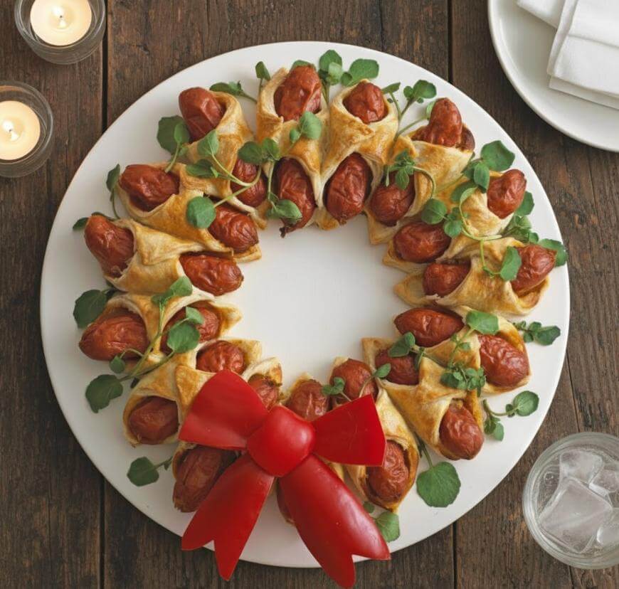 A round white platter with mini pigs in a blanket arranged in a wreath shape, garnished with fresh greens and topped with a red bell pepper bow. The platter sits on a wooden table with candles and napkins nearby.